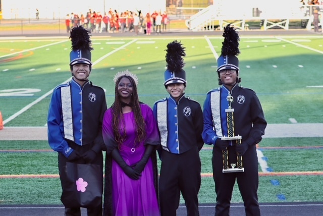 Four people stand on a football field, two in marching band uniforms and one in a purple dress, holding a trophy.