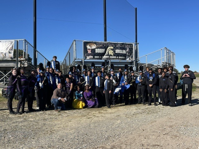 Group of band members standing in front of a metal structure with a banner, some seated and some standing.