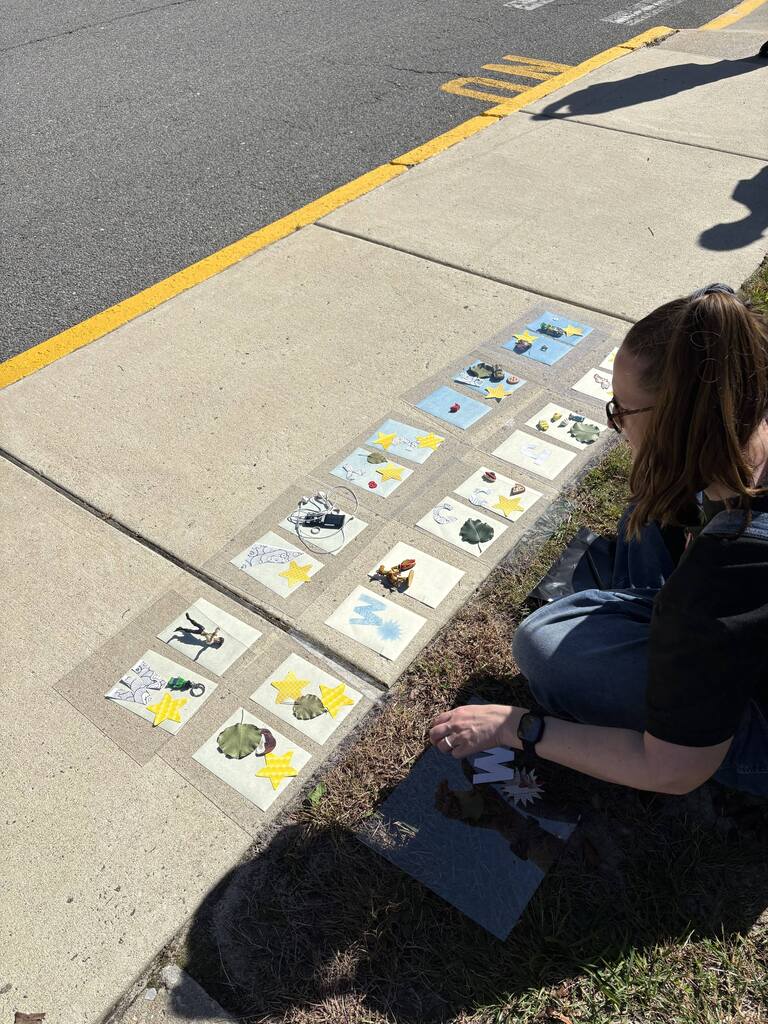 Woman sits on curb near street, with squares of paper on the sidewalk.