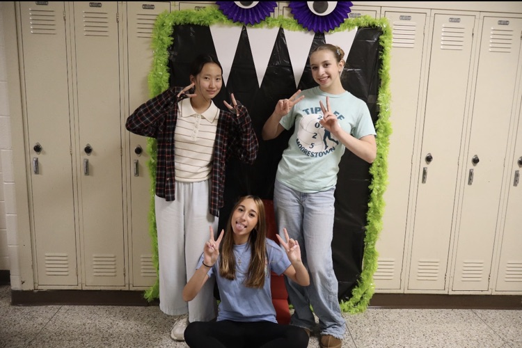 Three young girls pose for a photo in a hallway with lockers in the background.