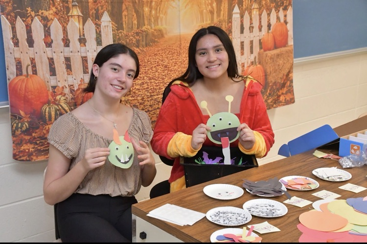Two girls hold up cut-out paper bugs at a table with paper plates, paper, and art supplies.