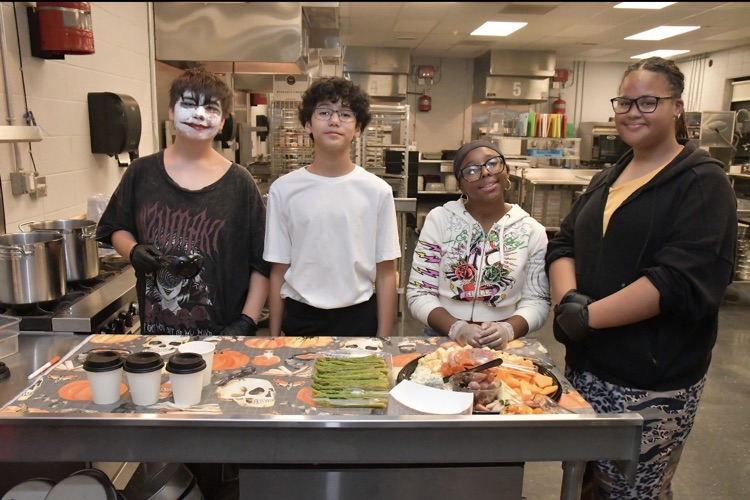 Four people in a kitchen with food and drinks on a table. One person has face paint.