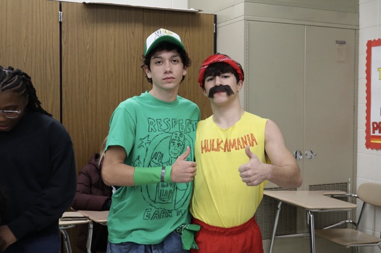 Three young males, two dressed in novelty costumes, stand in a classroom. One gestures thumbs-up.