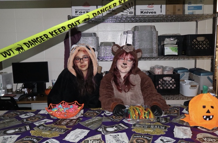 Two people in costumes sitting at a table with food and decorations; a pumpkin and a caution tape.