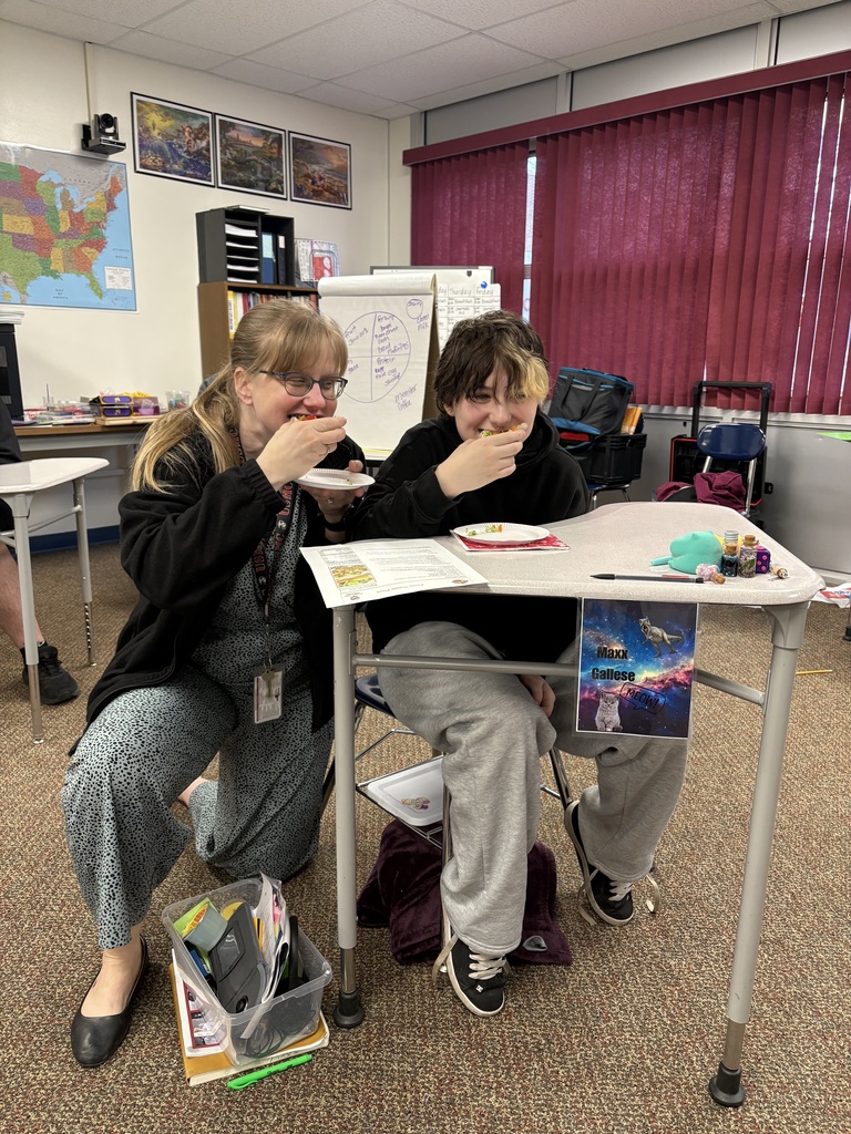 Two people enjoying pizza sitting at a desk in a classroom.