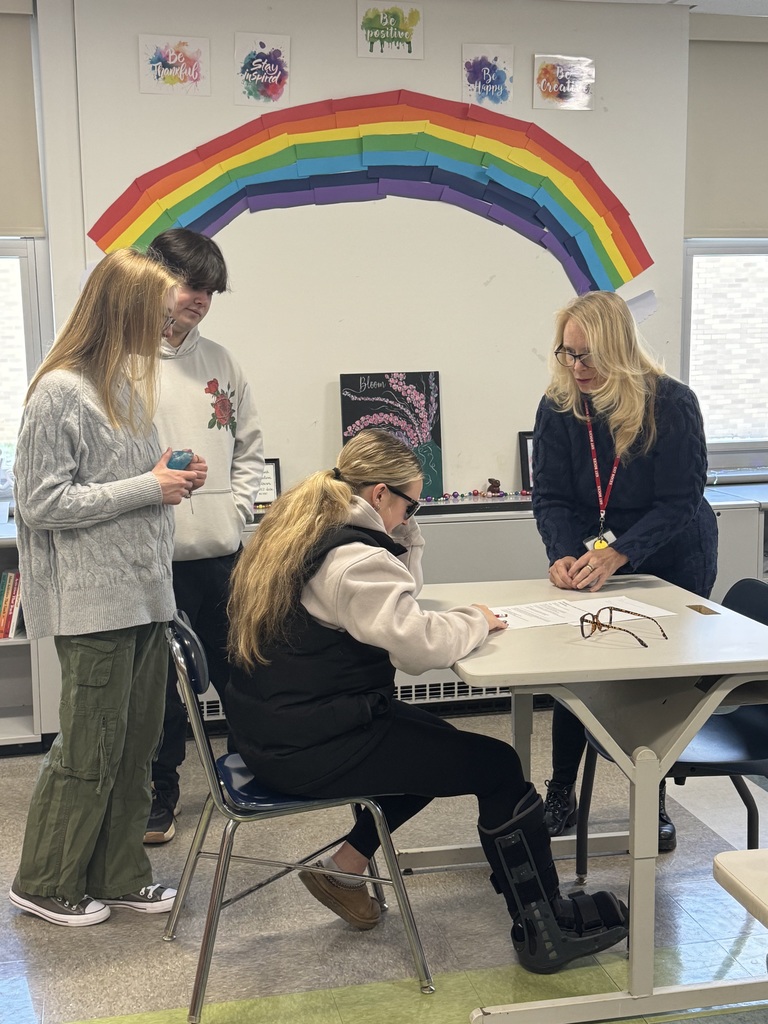 A teacher with three students, one of the students is sitting at a desk trying to read with glasses that make the page blurry.