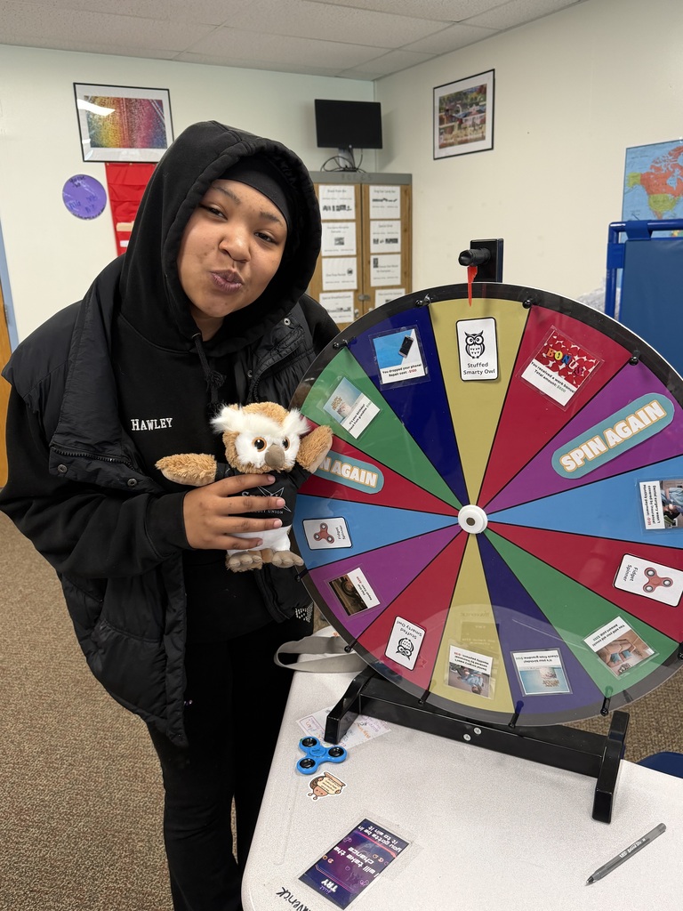 One student posing at a wheel to spin holding a stuffed owl.