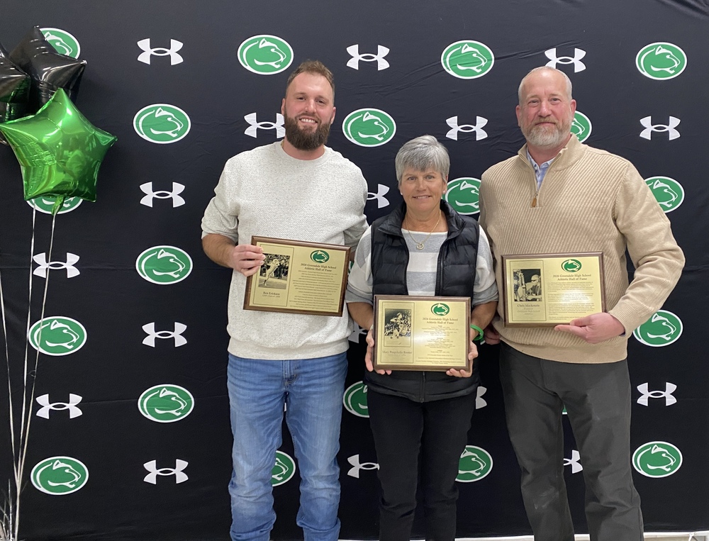 Photo of one woman and two men standing in front of a black and white backstop holding plaques announcing them as Hall of Fame Inductees