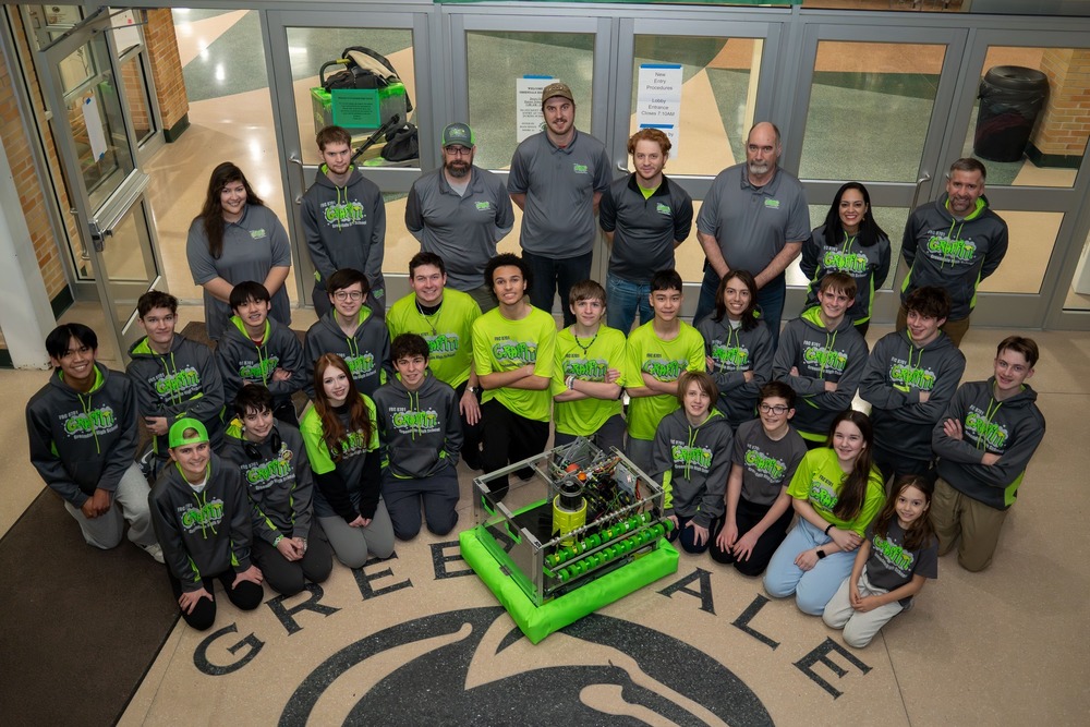 Students in school foyer wearing GRAFFITI robotics team shirts and posing with their robot