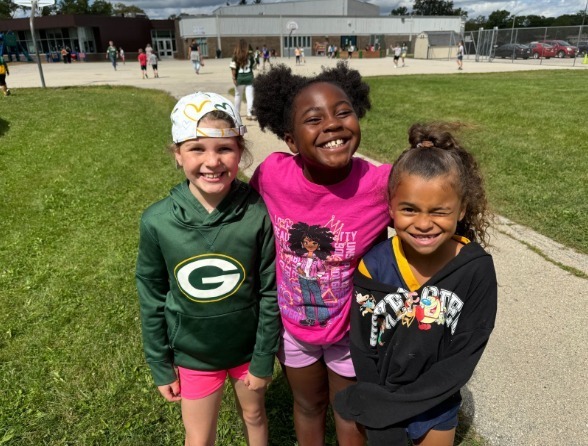 Three students standing on the grass smiling for the camera with the school in the background.