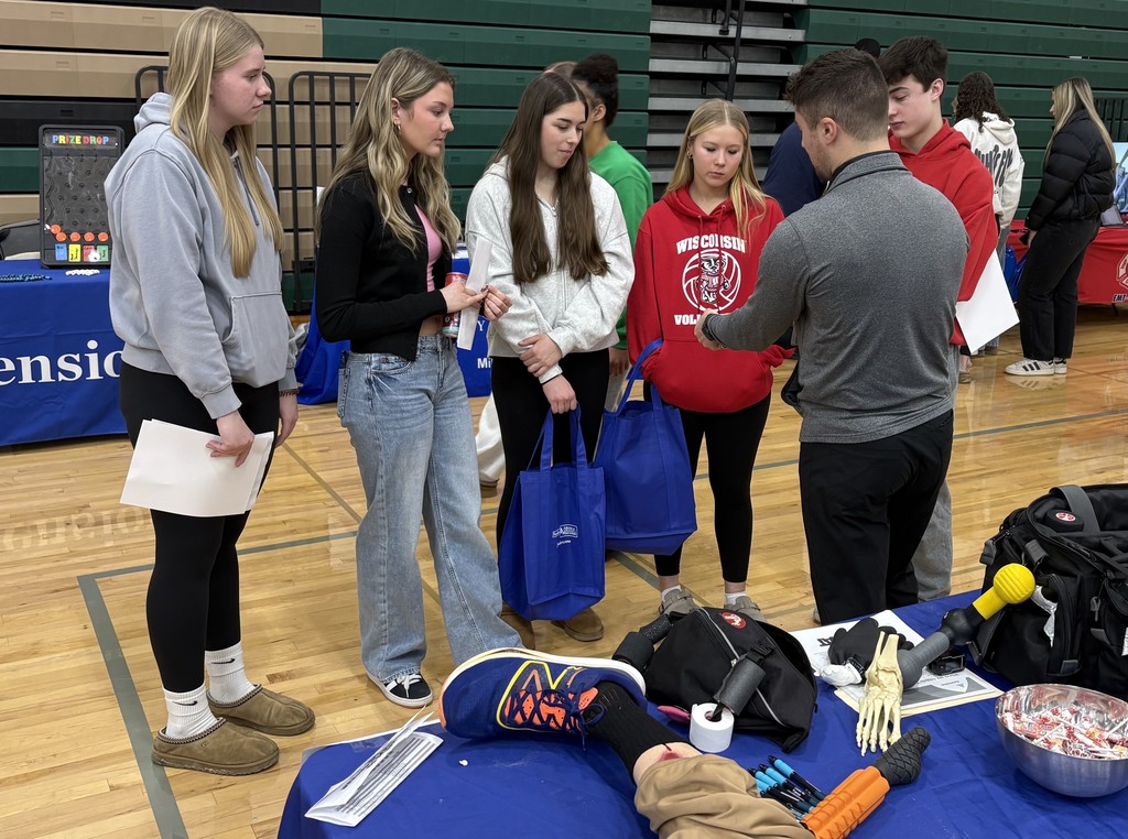 A group of people gathers around an informational booth in a gym, with a table displaying various items like a skeleton hand model and foam rollers.