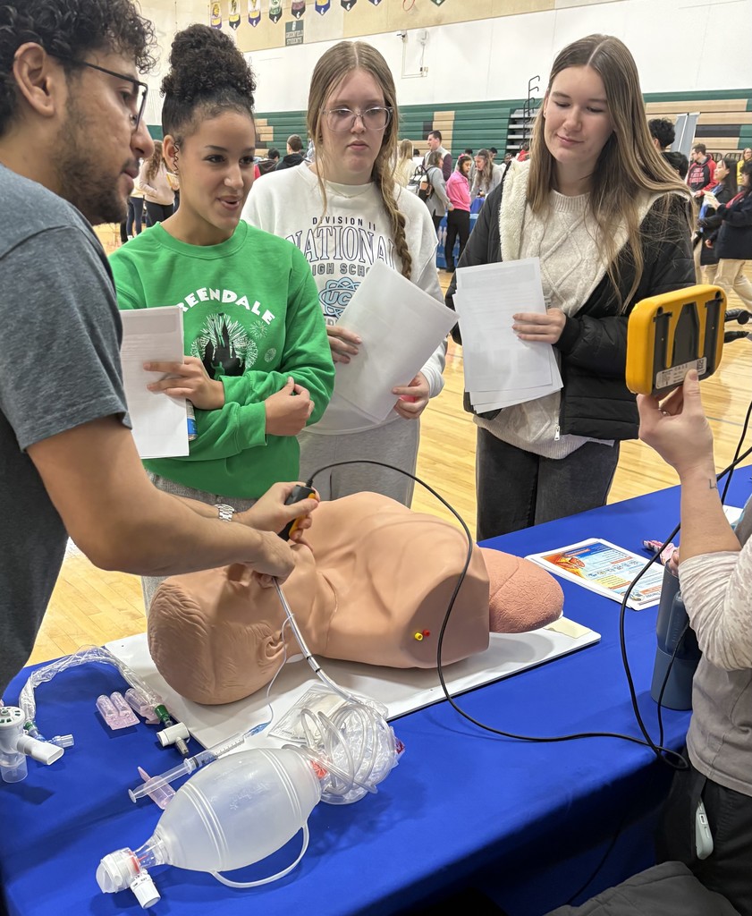 A group of people observing a medical demonstration on a mannequin in a gymnasium.