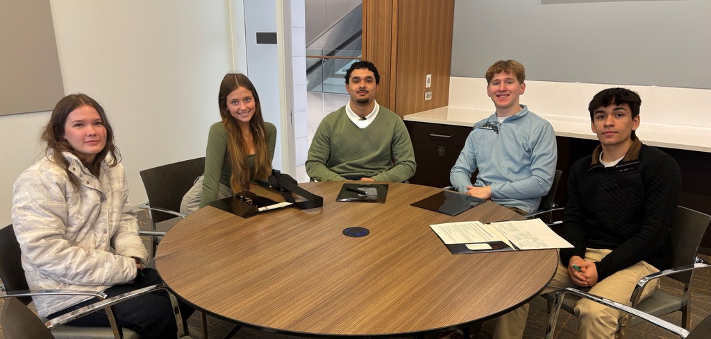 Five students at seated around a wooden table smiling for the camera