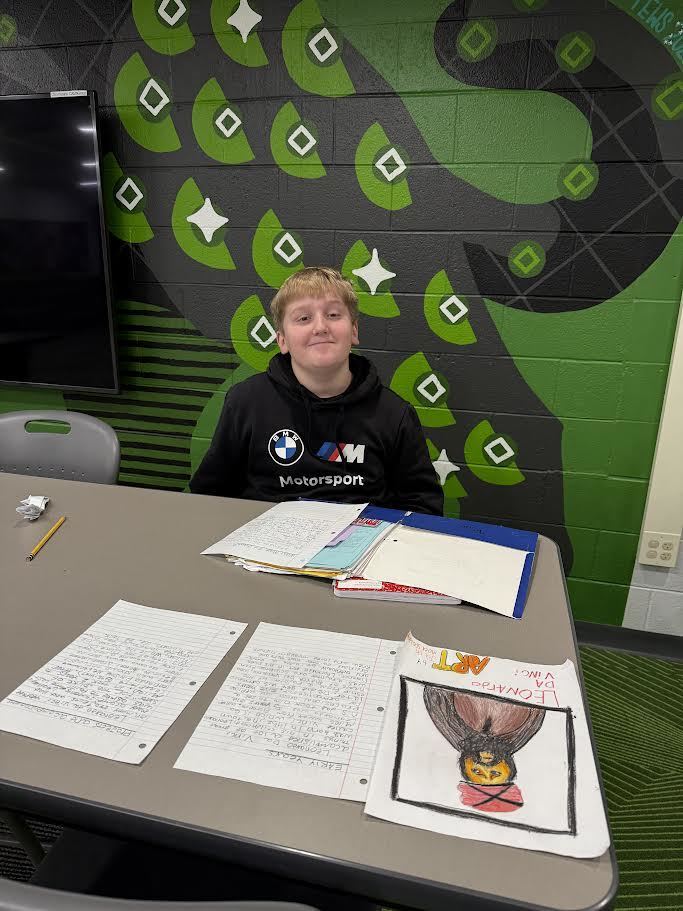 Fifth grade boy posing at desk with his ELA project spread out in front of him.