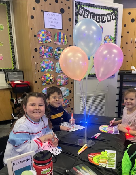 Three 5K students seated at a table working with markers in a 100th day of school celebration activity.