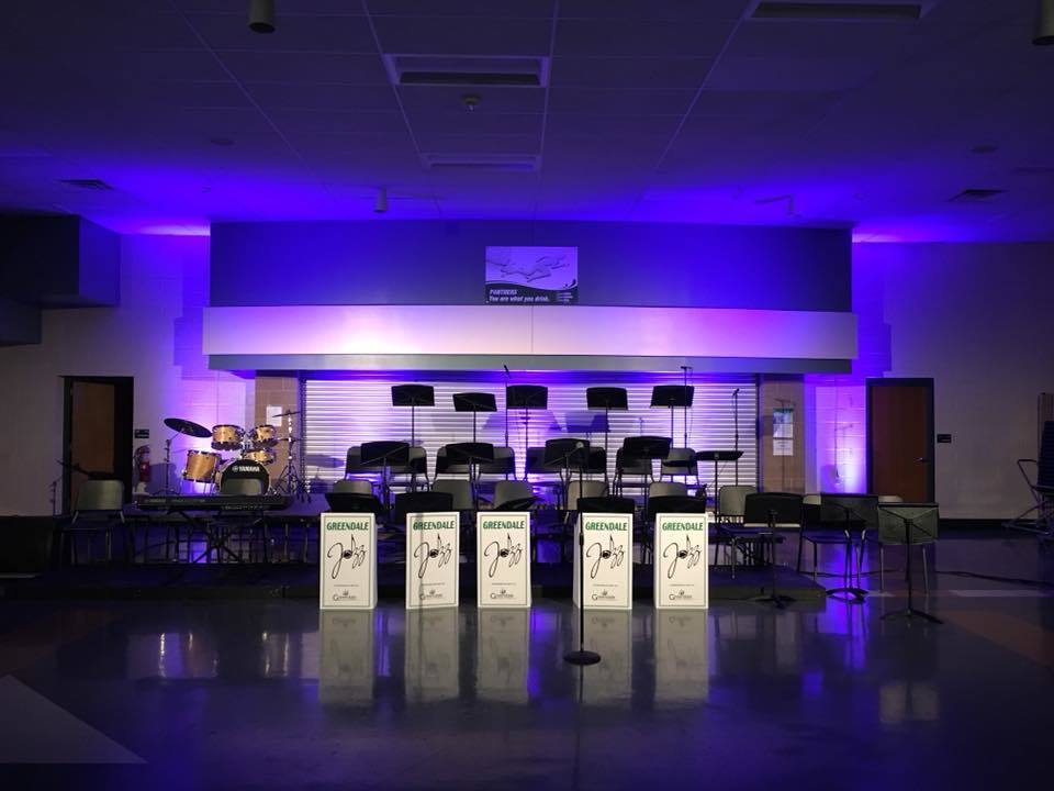 A stage set with music stands labeled "Greendale Jazz" and chairs, illuminated by purple lighting.
