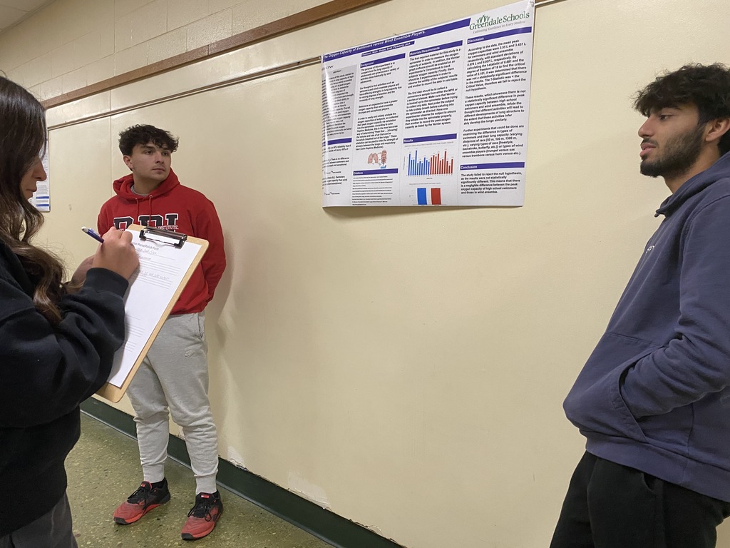 Three people in a hallway, two observing a research poster on the wall, and one writing on a clipboard.