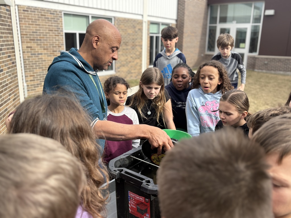 A man in a teal hoodie shows children a water-filled container outdoors.