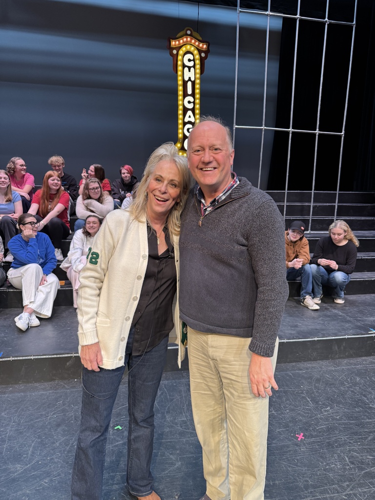 A smiling man and woman standing on a stage in front of a "CHICAGO" sign with a group of seated people in the background.