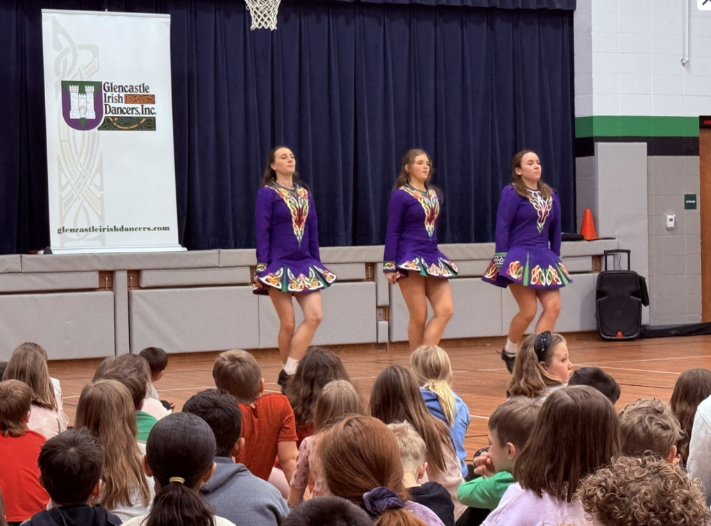 Three Irish Dancers performing together in front of a school assembly.