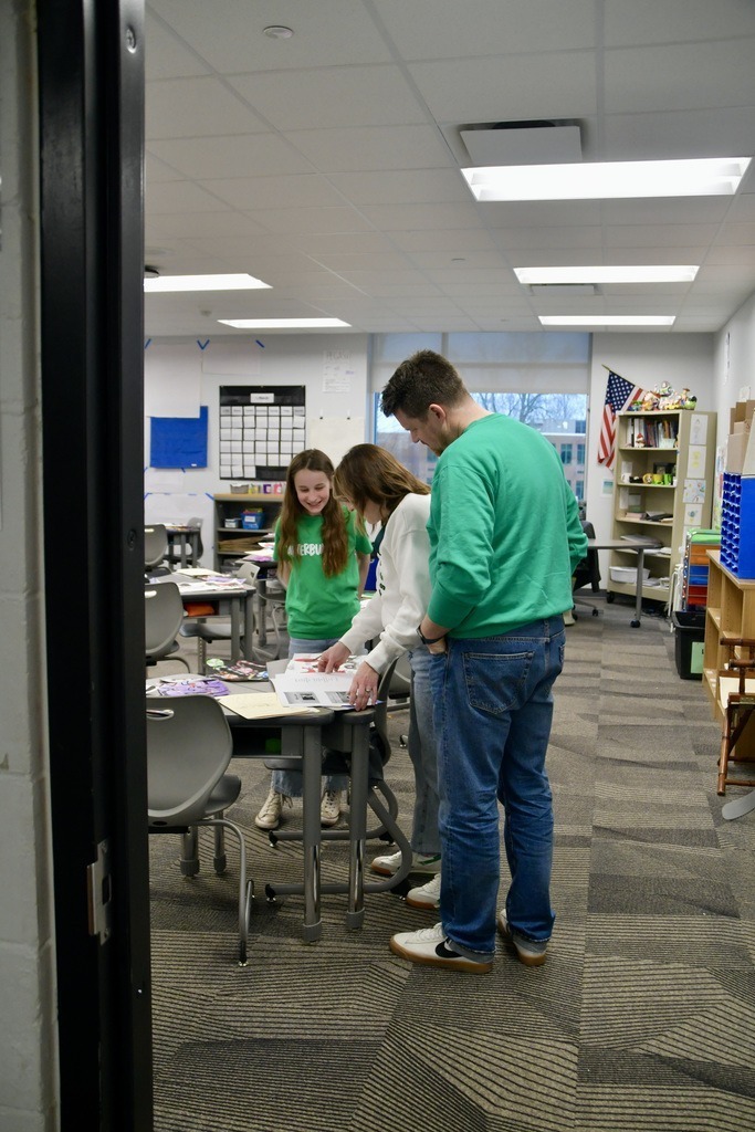 Three people in a classroom look at papers on a table under fluorescent lights.
