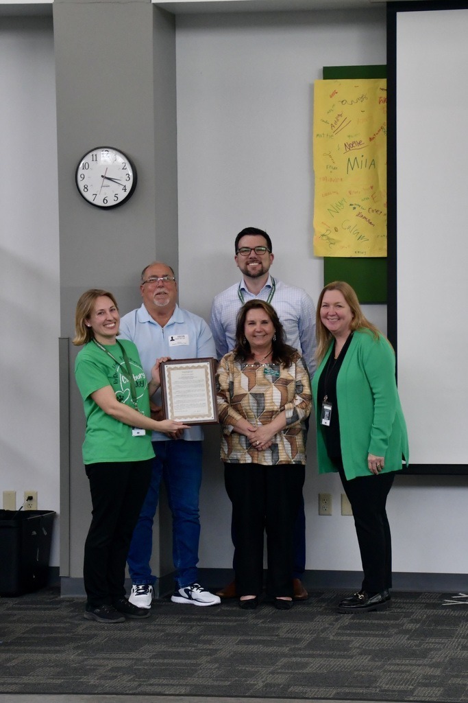 A group of five people smiling indoors, one holding a framed certificate.