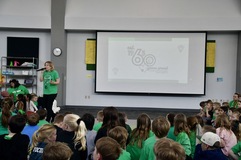 A school assembly with children in green shirts and a woman speaking into a microphone in front of a screen showing "60years proud."