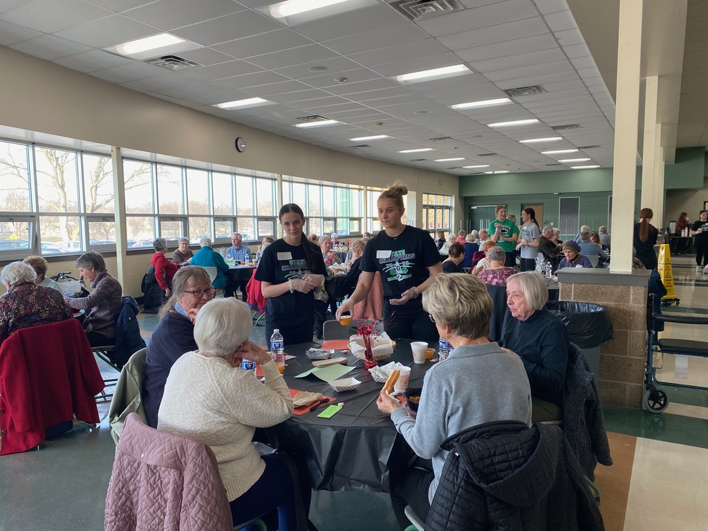 A community gathering with people seated around tables in a bright room, with staff serving food.