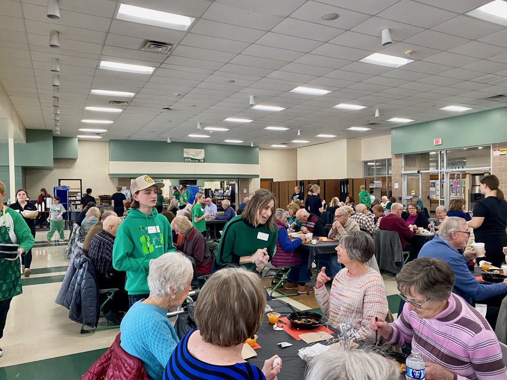 A community gathering with people seated around tables in a bright room, with staff serving food.
