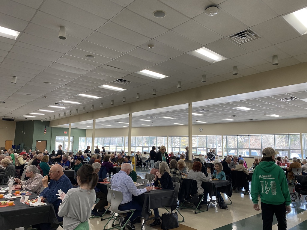 A community gathering with people seated around tables in a bright room, with staff serving food.