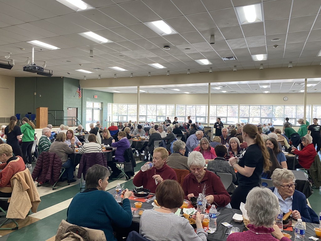 A community gathering with people seated around tables in a bright room, with staff serving food.