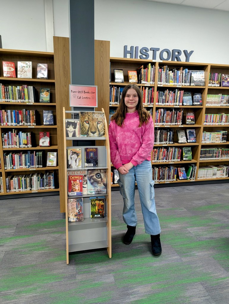 A person stands beside a cat-themed book display in a library with shelves labeled "HISTORY" in the background.