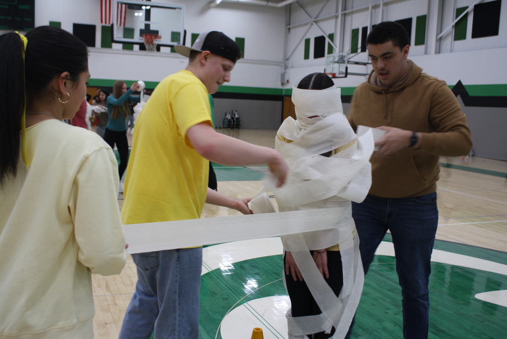 Four people in a gym, three wrapping the fourth in toilet paper.