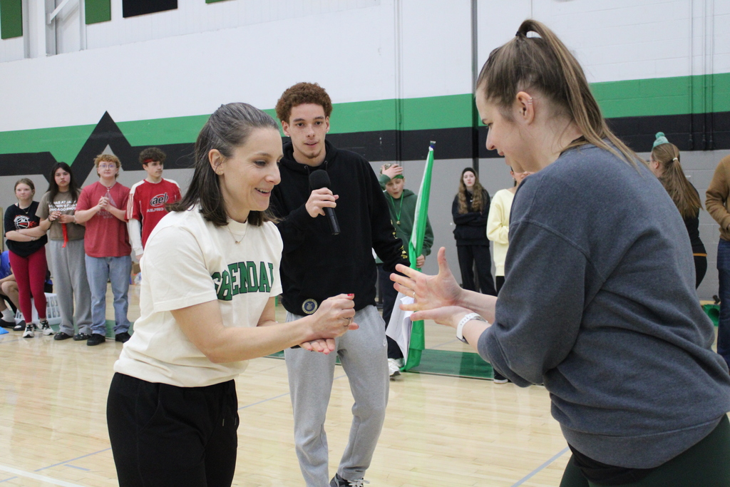 Two women play rock-paper-scissors, watched by a young man with a microphone in a gym.