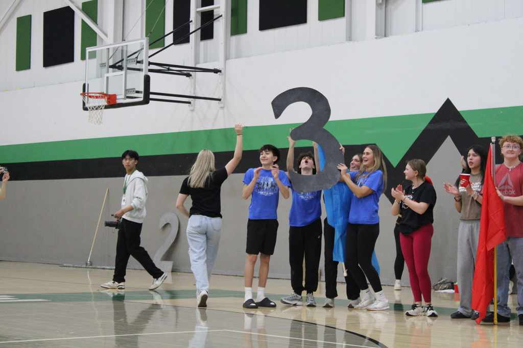 A group of young people in a gym, holding a large number "3", with a basketball hoop above them.