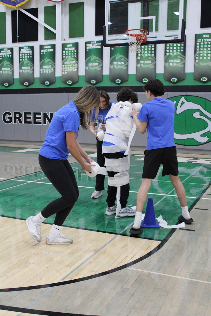 Four people in a gym, three wrapping the fourth in toilet paper.