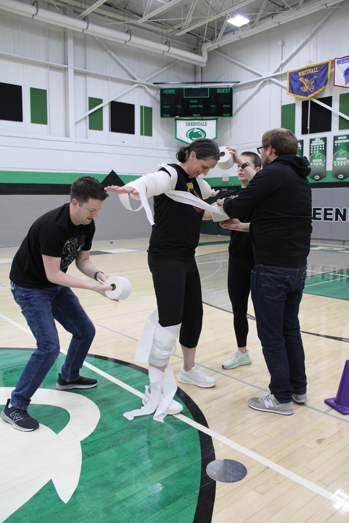 Four people in a gym, three wrapping the fourth in toilet paper.