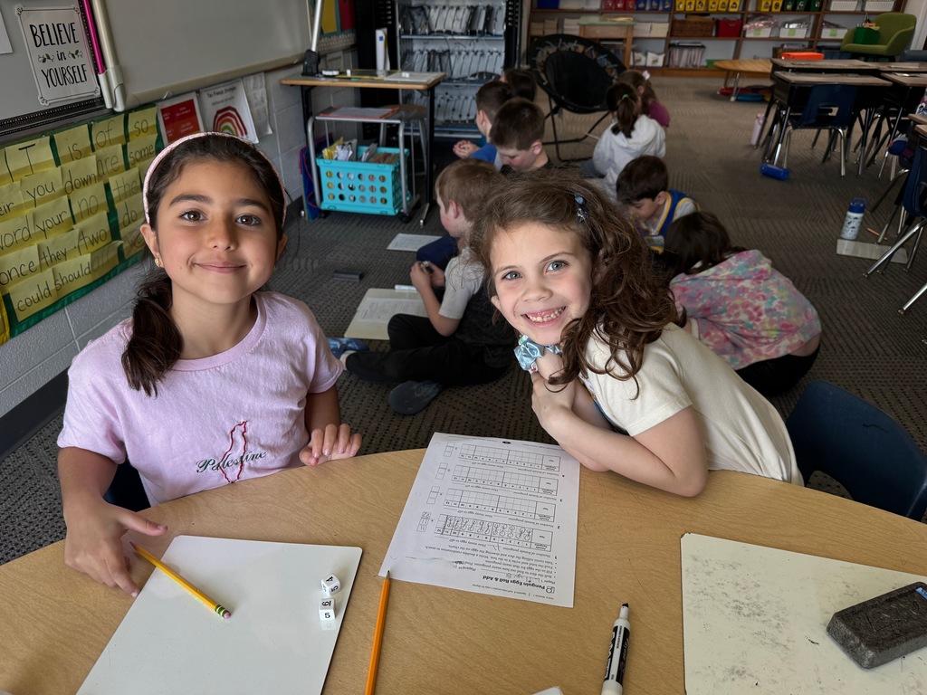 Two children sitting at a table doing math worksheet with a dice and markers.