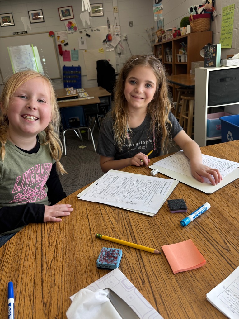Two children sitting at a table doing math worksheet with a dice and markers.