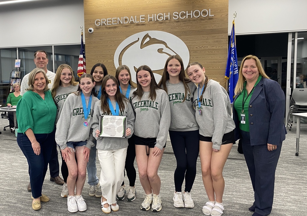 A group of young women in grey sweatshirts labeled "Greendale Poms" stands with two adults in front of a wall with the "Greendale High School" emblem.