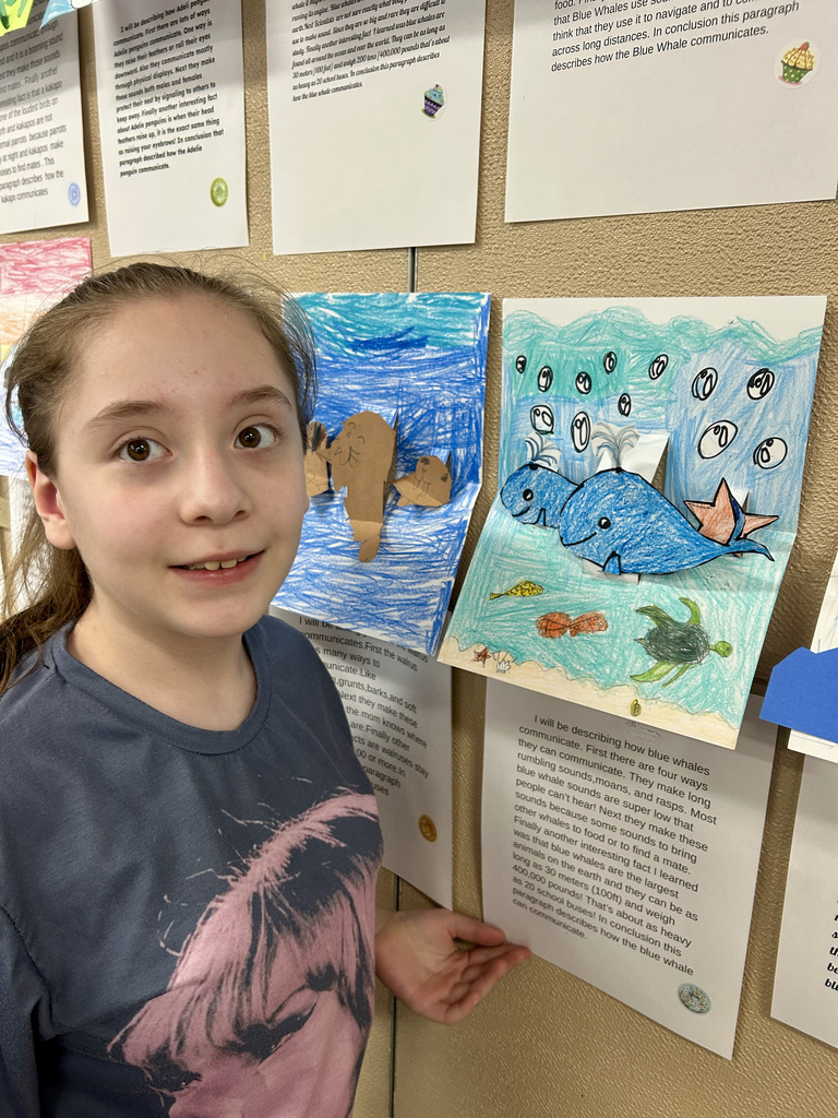 Girl standing by a wall of posted projects on animals with her hand up by hers..