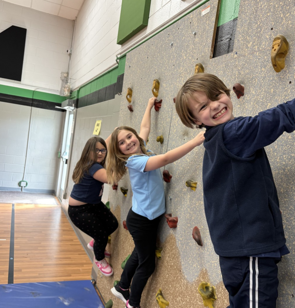 Three smiling students climbing on the school's climbing wall in PE.