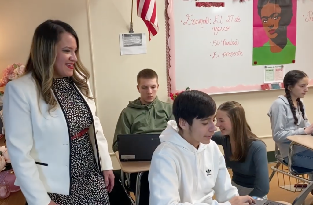 Teacher in white jacket speaking to a student and three other students in the background are working