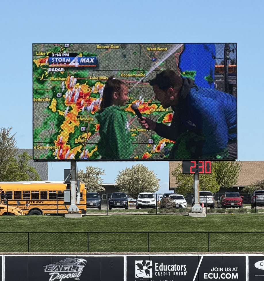 Student on big screen talking with TMJ4 staffer during weather show on baseball field trip.