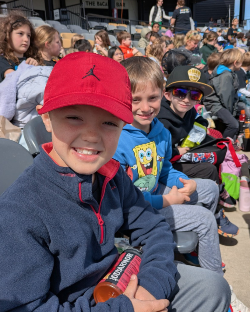 Students smiling and seated in the stands at Franklin Field during a baseball field trip.