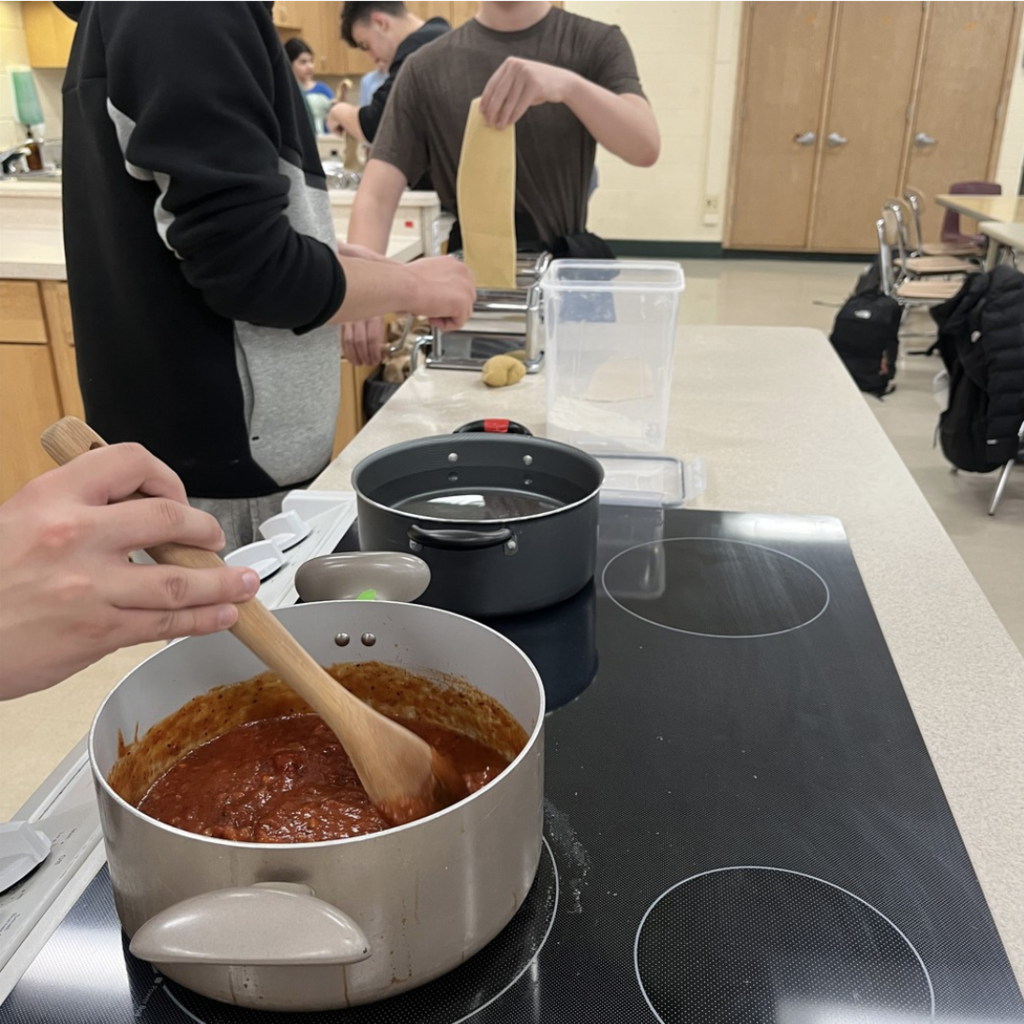 Kitchen scene with students rolling out homemade pasta and a kettle of red sauce being stirred on the stove.