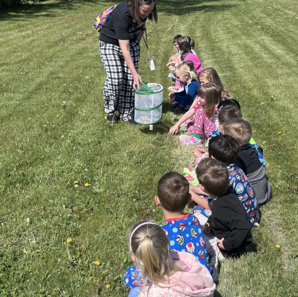 4K students sit on the grass watching a teacher open a net butterfly house.