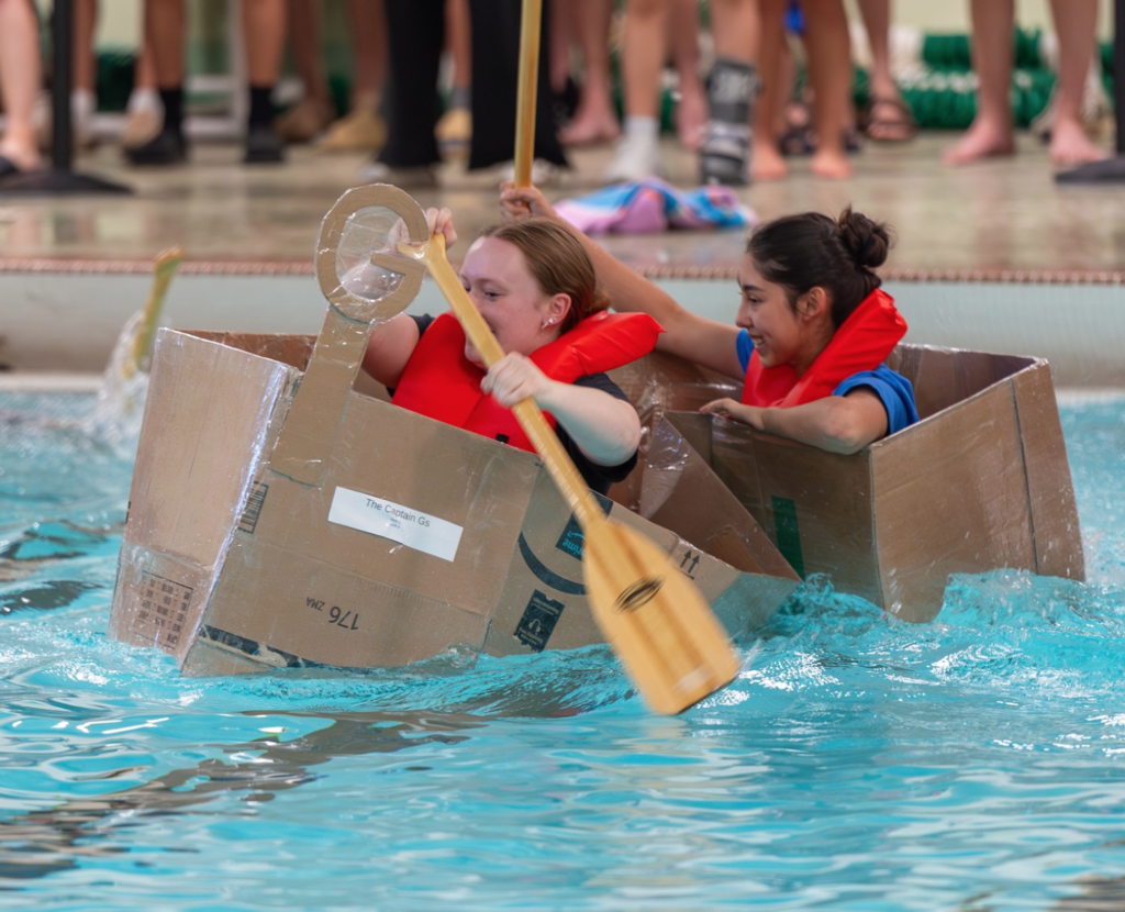 Two students in life jackets paddling inside a cardboard box boat in the pool for the Physics Regatta.