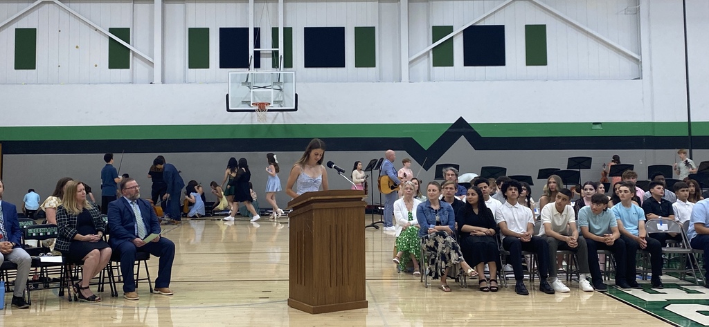 A school event in a gymnasium with a youth speaking at the podium, other adults in seats and students seated and musicians standing in the background.