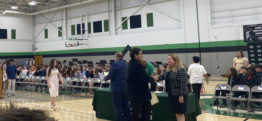 A school event in a gymnasium with students receiving a certificate and shaking hands with adults and other students seated in the background.